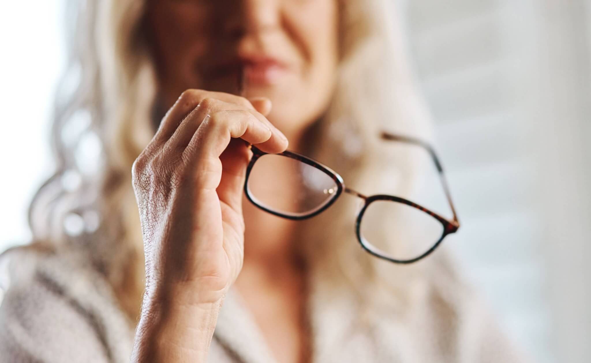 Nahaufnahme einer Hand, die eine Brille hält, im Hintergrund eine unscharfe Person.