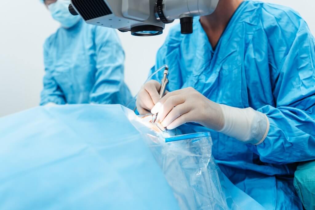 Chirurg in blauer OP-Kleidung führt Augenoperation unter Mikroskop durch.