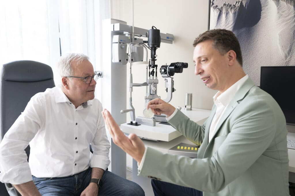 Augenarzt berät älteren Mann in Praxis, zeigt auf Augenuntersuchungsgerät, helle Umgebung.