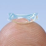 Nahaufnahme einer implantierbaren Kontaktlinse auf einer Fingerspitze vor blauem Hintergrund.