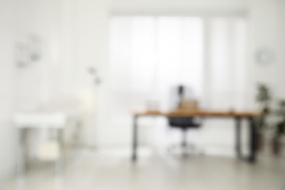 Verschwommenes Bild eines hellen, minimalistischen Büros mit Schreibtisch und Stuhl vor einem Fenster.